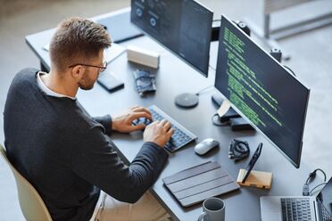 Ein Mitarbeiter arbeitet an einem Schreibtisch mit zwei Monitoren, auf denen Code für ein Dokumentenmanagement-System zu sehen ist.